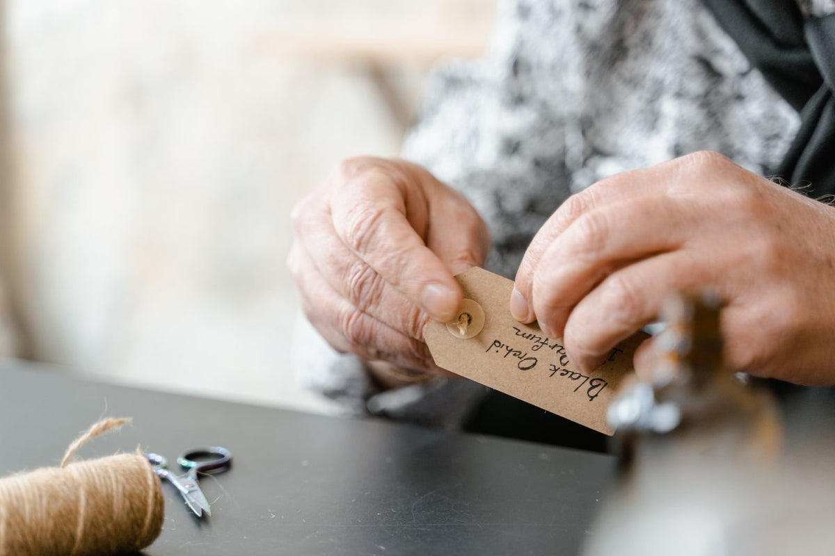 Mains d'artisane fixant une étiquette kraft manuscrite sur une création, bobine de ficelle et ciseaux posés sur l'établi