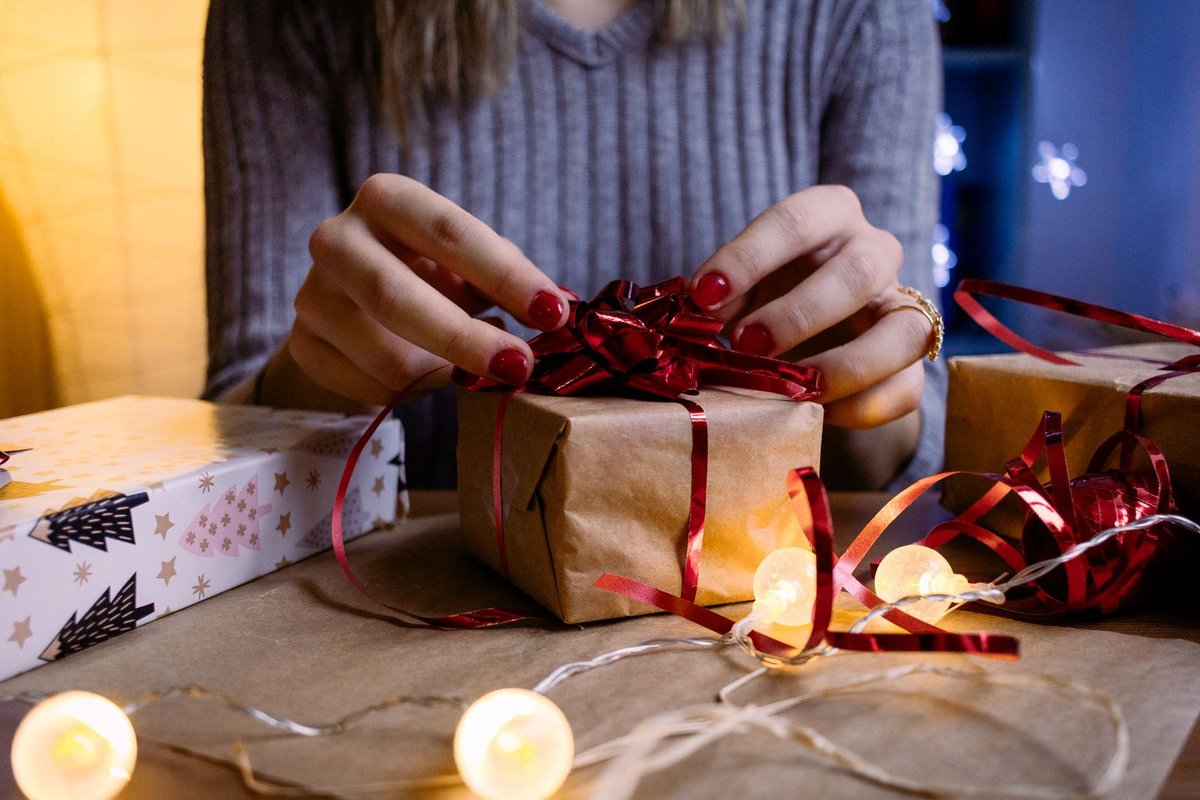 Mains nouant un ruban rouge sur un colis en papier kraft, guirlande lumineuse allumée sur la table