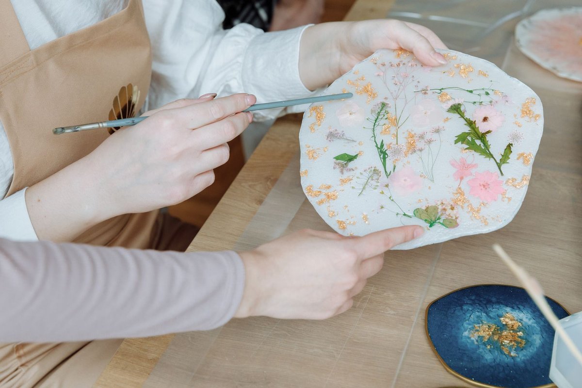 Mains présentant un dessous de verre en résine aux fleurs séchées et éclats de feuille d'or, pièce bleu nuit posée sur la table